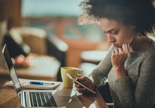 A woman wearing a brown jersey sits at her desk looking at her cellphone with her computer behind it, a notepad and pence and a yellow coffee mug to her right.