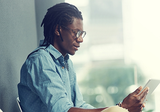 A man with short dreadlocks and a blue shirt sitting a table accessing his bank account via his mobile.