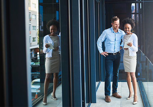 Full length image of two colleagues - a man and a woman - standing on the balcony outside their office looking at the screen of a mobile device. The street scene is reflected in the office window.